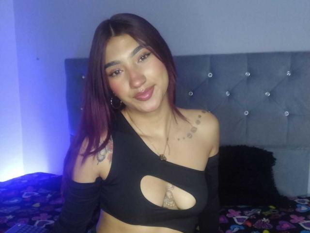 NemesisSinner with dark purple hair, black cut-out top showing tits, tattoo on shoulder, nose ring, and black sleeves, smiling in a bedroom with blue lighting.