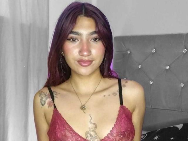 NemesisSinner with purple hair, red lace bra, tattoos on shoulders and chest, small tits, necklace, pouty lips, standing in front of a grey tufted headboard.