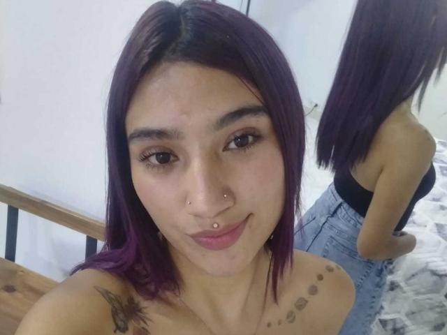NemesisSinner with purple hair, nose piercings, and a tattoo on her shoulder, smiling at the camera, wearing a black top and high-waisted jeans, reflected in a mirror.