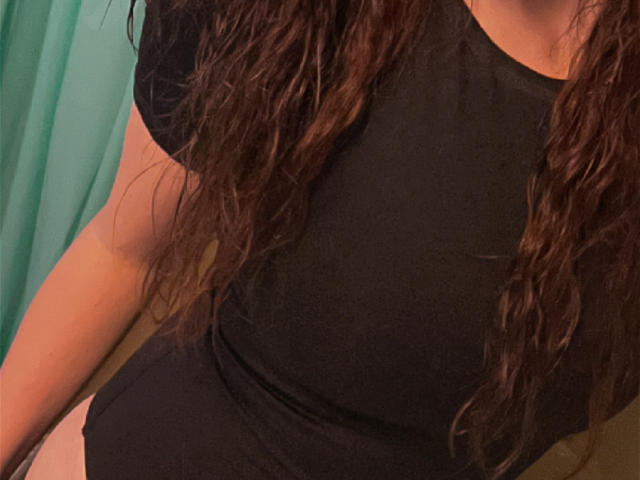 La parte superior del cuerpo de TinaLocket, con cabello castaño largo y ondulado, lleva una camiseta negra que muestra sus senos y una pequeña franja de su ropa interior blanca. Su brazo se ve parcialmente..