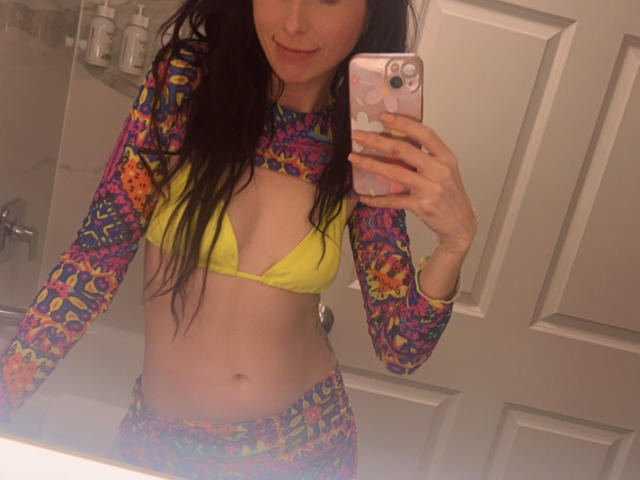 TinaLocket, con lunghi capelli scuri, indossa un top da bikini giallo e un top lungo colorato con motivi psichedelici, abbinato a pantaloni coordinati, e sta scattando un selfie allo specchio in un bagno..