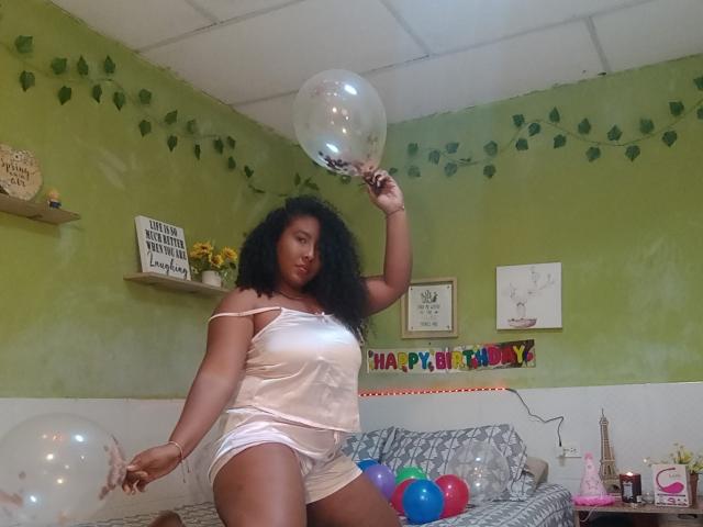 CaroldelaTorre with curly black hair, dark skin, and curvy body, wearing a white satin off-shoulder top and shorts, holding a clear balloon, surrounded by colorful balloons.