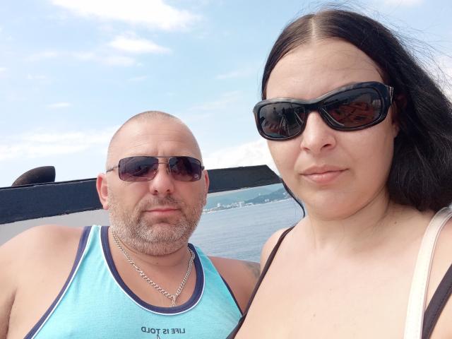 GretaTamar with bald, bearded man in blue tank top and sunglasses, and GretaTamar with black hair, brown eyes, black sunglasses, and cleavage in white top. Boats in background.