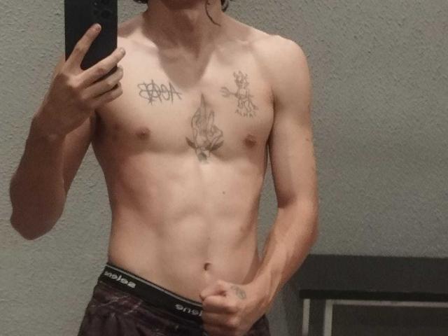 Hiesoka's slim, toned torso with small nipples, tattoos on chest and upper arms, wearing dark pants with "PUMA" waistband, holding phone in mirror selfie.