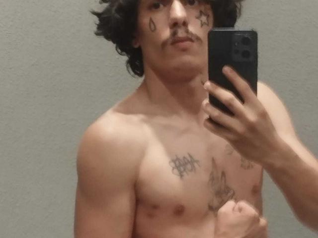 Hiesoka, shirtless with dark curly hair and a mustache, holds a phone in a mirror selfie, showing tattoos on his chest and neck, and a flaccid nipple.