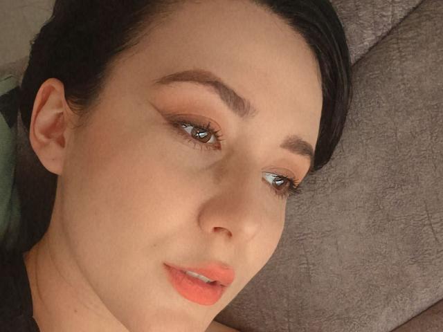 MabyLegrand has fair skin, dark eyebrows, shiny blue eyes, and orange lipstick. She's lying on a grey textured surface with her head slightly tilted. Her black hair is partially visible.