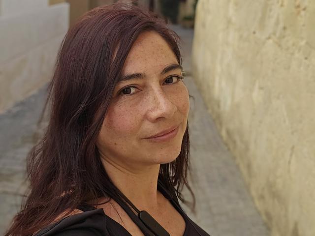 AlessiaRed a la peau constellée de taches de rousseur, des yeux marron et de longs cheveux bruns foncés. Elle sourit légèrement, elle porte un haut noir, et un mur jaune texturé se trouve en arrière-plan..