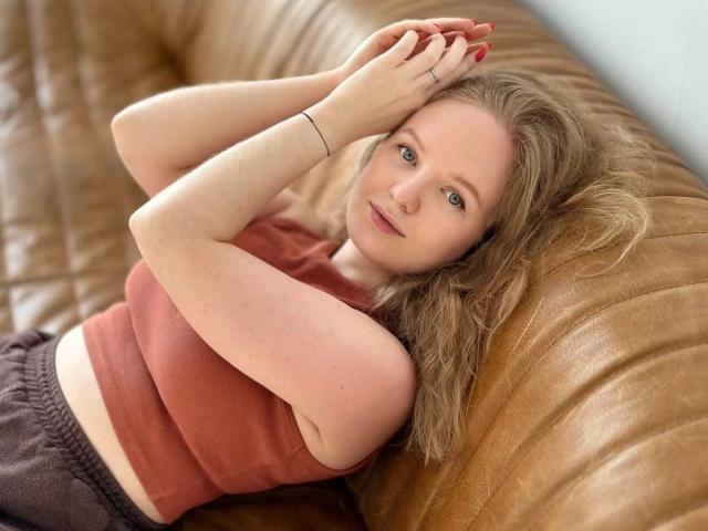 SunnyBunny-hot with wavy blonde hair, red crop top, and brown pants, lying on a tan leather couch, hands on her head, looking at the camera, smiling.