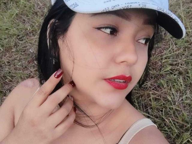 KarlyShiine with red lipstick, black eyeliner, and red nail polish, wearing a white cap, touching her neck, brown hair, and a beige top, standing on grass.