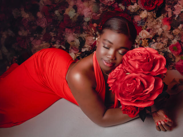 KarlieMoss, à la peau foncée, allongée sur le côté, portant une robe rouge, tenant de grandes roses rouges, les yeux fermés, peau lisse, ongles rouges, fond floral, sexy, attirante, sourire doux..