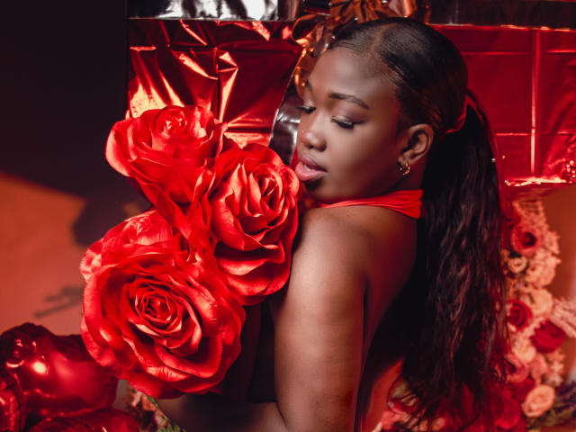 KarlieMoss with dark skin, long wavy hair in a red ponytail, holding shiny red roses, wearing a red halter top, looking down with closed eyes, red background.