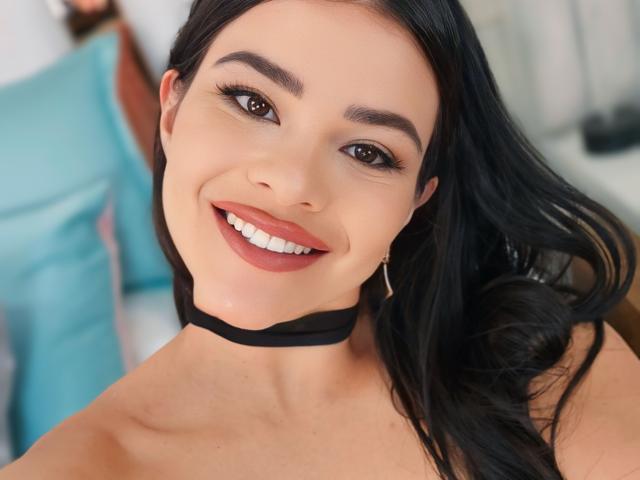 PaulinaValley with black hair, brown eyes, and a smile, wearing a black choker, showing her tits, and a red lipstick. She's also wearing small hoop earrings.