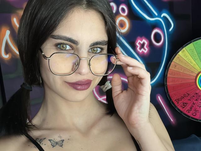 AngelCharmy with green eyes, dark pigtails, and glasses, holding her glasses, has a butterfly tattoo on her chest and wears purple lipstick. Neon lights and a wheel are in background.