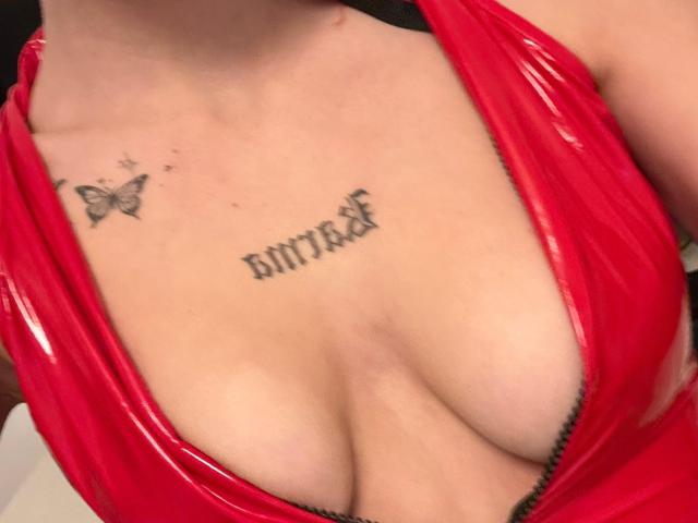 AngelCharmy's red latex dress reveals her big, shiny, cleavage-filled tits. She has a butterfly tattoo on the left and "Bitter" tattooed below it.