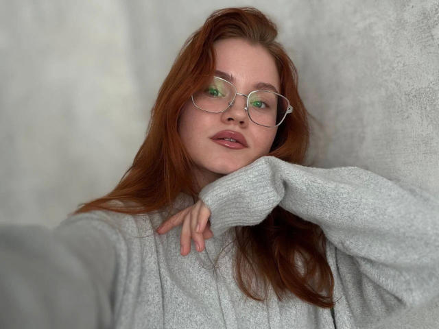 Lumuon-hot, met rood haar, groene bril en een grijze trui, heeft haar hand onder haar kin, kijkt naar de camera met een lichte glimlach, en laat haar roze lippen en sproeten zien..