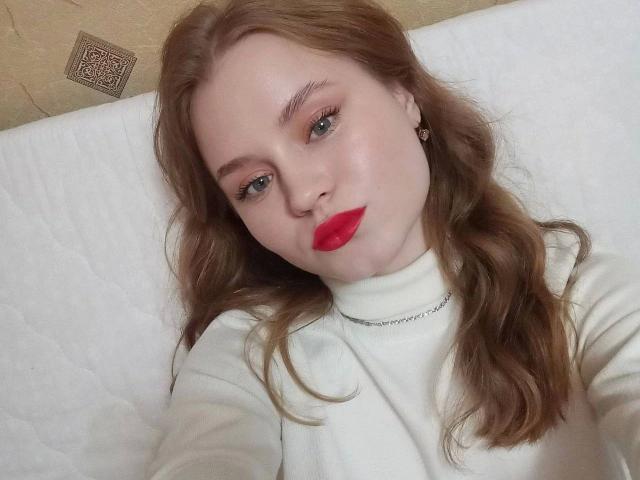 Een vrouw met golvend bruin haar, rode lippenstift en een witte turtleneck, liggend op een wit, gewatteerd oppervlak, draagt kleine, hartvormige oorbellen en kijkt met een lichte glimlach naar de camera..