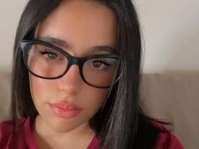 AlexaBabeZ has tan skin, dark straight hair, black glasses, and pink lips. She's wearing a red shirt with a serious expression. Her tits are not visible.