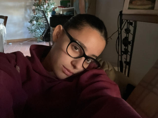 AlexaBabeZ with glasses, dark hair in a ponytail, wearing a red hoodie, resting her head on her arm, looking at the camera with a slight smile.
