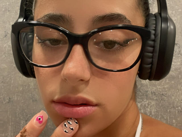 AlexaBabeZ with glasses, black headphones, and pink nail with white and black pattern, touching her lips. Her skin is tan, and she has brown eyes and pink lips.