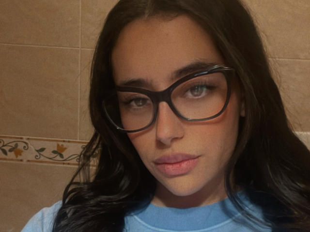 AlexaBabeZ with glasses, dark wavy hair, light pink lipstick, blue shirt, serious expression, slightly flushed face, green eyes, tan skin, tile wall background.