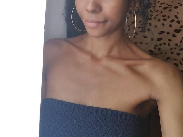Mouniah with dark skin, curly black hair, small hoop earrings, and a navy blue, strapless, textured top, showing her collarbones and a subtle smile.