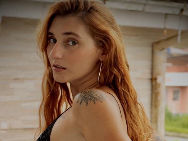 Melissadry with long red hair, hoop earrings, and a sun tattoo on her right shoulder, looking over her shoulder with a slight smile. She's wearing a black lace top.