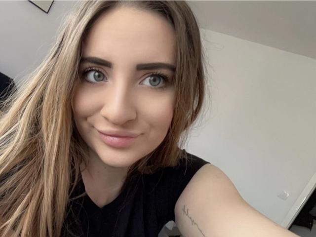 DakotaGold has blue eyes, long brown hair, and a slight smile. She's wearing a black sleeveless top, showing a tattoo on her right arm.