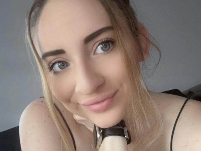 DakotaGold with blonde hair, blue eyes, and a slight smile, wearing a black strap top, resting her chin on a white dildo, looking at the camera.