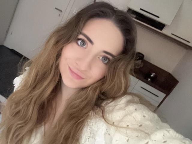 DakotaGold, con lunghi capelli castani ondulati, occhi azzurri e un leggero sorriso, indossa una maglia bianca con una texture particolare, in un selfie. Il suo seno è piccolo e i suoi glutei non sono visibili..