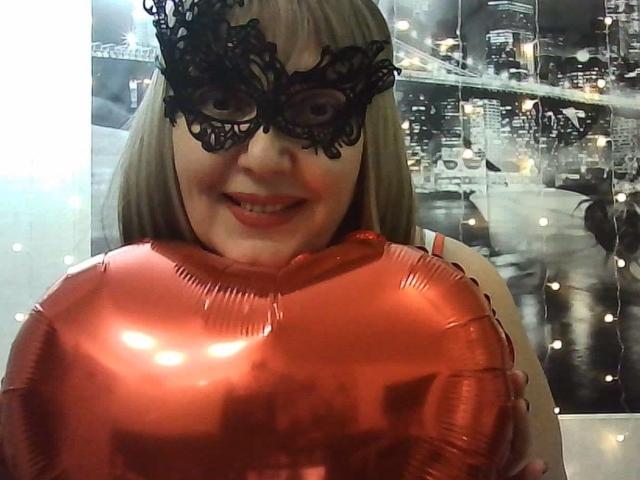 CarlySweetie with a black lace mask, smiling, holding shiny red balloons, showing her tits, blonde hair, and cute face.