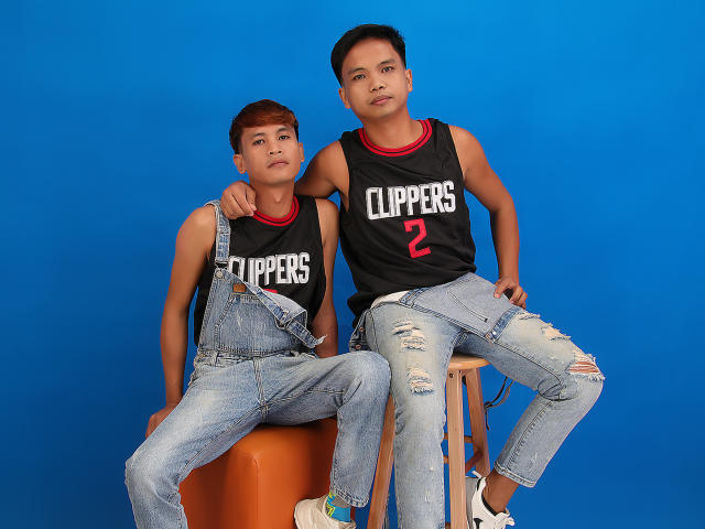Two ExtraService69MenX in black "CUPPERS" tank tops and ripped jeans, sitting on stools against a blue background. One with short brown hair, the other with short black hair.