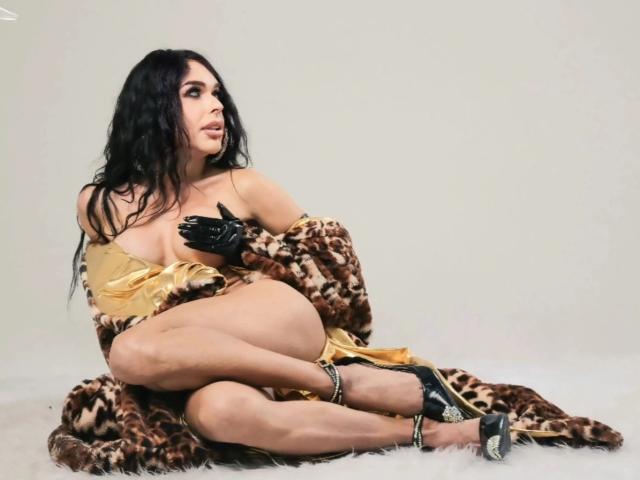 Jhovanna with black wavy hair, leopard fur robe, gold fabric, black gloves, and black high heels, sitting with crossed legs, showing her tits, looking to the side.