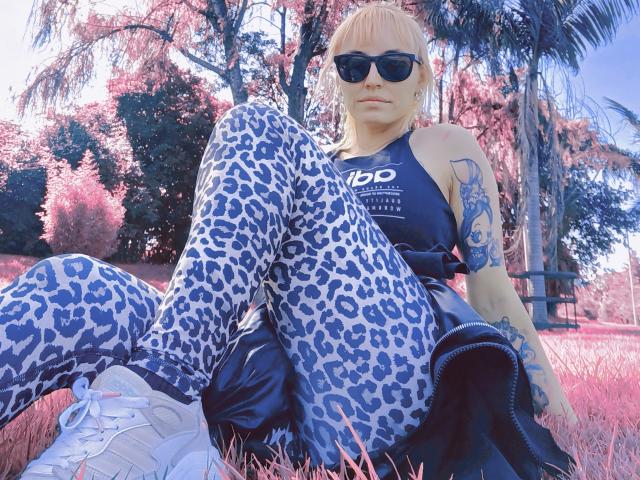 SaraleePoison with bleached blonde hair, black sunglasses, leopard print pants, white sneakers, and black top, sitting outdoors, showing tattoos, with a black jacket draped over her ass.