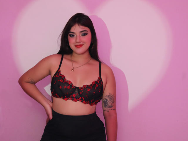 VictoriaSaens with red and black lace bra, showing her tits, black skirt, tattoo on right arm, pink heart shadow, long brown hair, red lipstick, smiling, hand on hip.