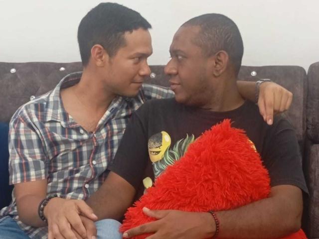 BillyAndMarkus, avec ses cheveux noirs courts, sa chemise à carreaux et son bracelet, est assis à côté de BillyAndMarkus, qui a la tête rasée, porte un t-shirt noir et est assis sur un coussin rouge moelleux, sur un canapé marron..