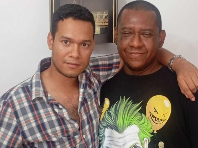 BillyAndMarkus, com cabelo preto curto, camisa xadrez e leve pelos no peito, está ao lado de BillyAndMarkus, com a cabeça raspada, camisa preta e estampa de balão do Coringa, sorrindo..
