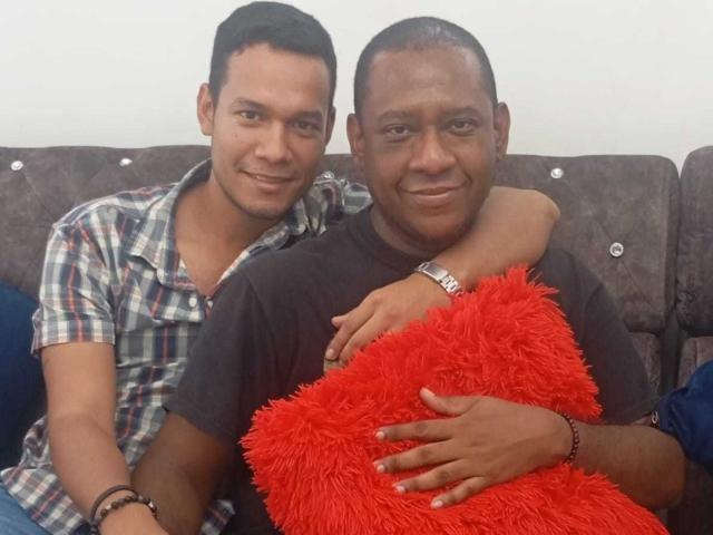 BillyAndMarkus (à esquerda), vestindo uma camisa xadrez, tem o braço ao redor de BillyAndMarkus (à direita), que está segurando um travesseiro vermelho fofo. Ambos estão sorrindo, sentados em um sofá cinza..