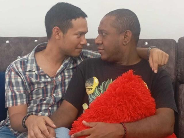 BillyAndMarkus, com cabelo preto curto e vestindo uma camisa xadrez, está sentado ao lado de BillyAndMarkus, com a cabeça raspada, vestindo uma camiseta preta e segurando um travesseiro vermelho felpudo. Eles estão em um sofá..