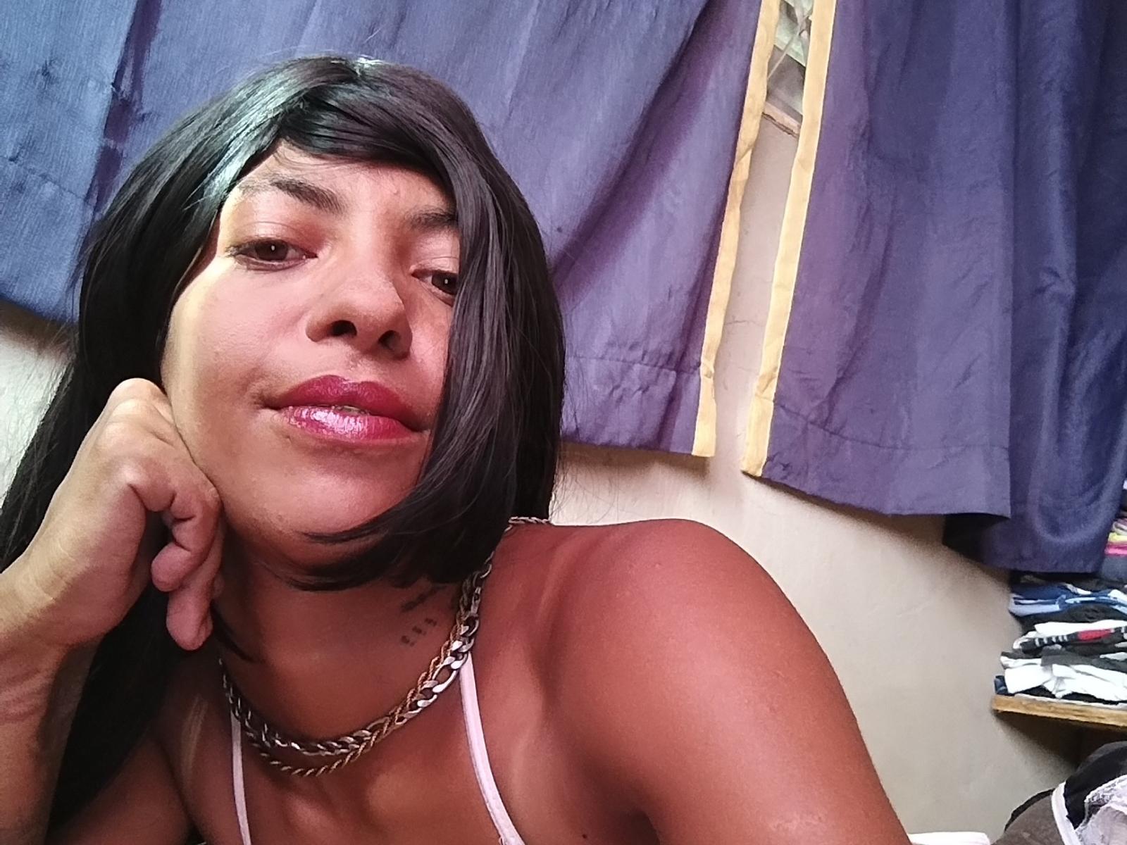 MissBloomy with glossy pink lips, short black hair, wearing a silver chain necklace, and a white halter top, resting her chin on her hand, smiling slightly.
