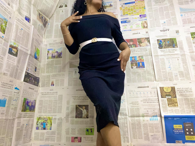 SophieSonyas with dark blue off-shoulder dress, white belt, and curly black hair, standing, hands on hips, showing her ass and tits, against newspaper-covered wall.