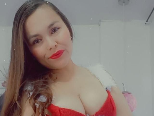 PinkyHotLover trägt roten Lippenstift, einen roten Spitzen-BH und hat braunes Haar. Sie zeigt ihre Brüste und trägt ein flauschiges, weißes Schulteraccessoire. Ihre Haut ist hell und sie lächelt..