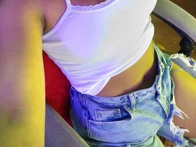 A camiseta branca de AfroditaKane revela seus pequenos seios, com calças jeans azuis rasgadas cobrindo suas nádegas. Sua piercing no umbigo roxa é visível. Ela está sentada em uma cadeira cinza..