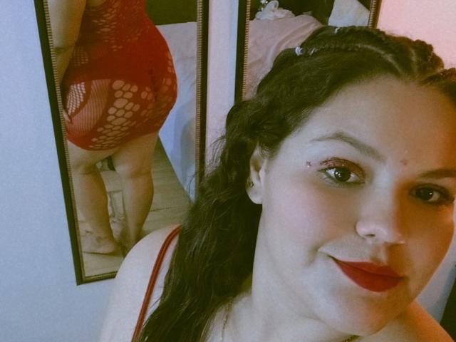 Gordita69 in red fishnet dress, showing her round ass in mirror selfie. She's smiling with red lipstick, dark braids, and a nose ring. Her tits are visible in dress.