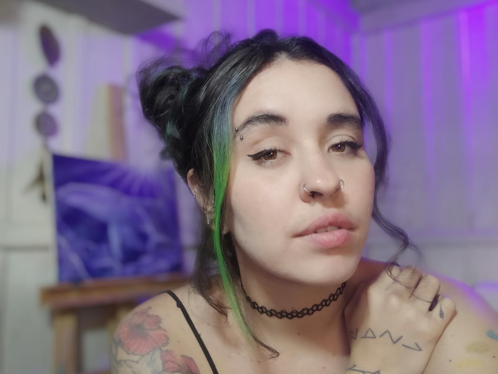 Sunyy with green-tinged black hair, nose ring, eyeliner, choker, tattoos, and pouty lips, showing off her tits and looking sexy. Blurry purple background, casual setting.