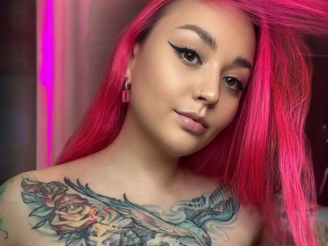 PinkHurricane-hot with bright pink hair, thick black eyeliner, tattoos of roses and birds on her chest, and a subtle smile, looking down with pink ear studs.
