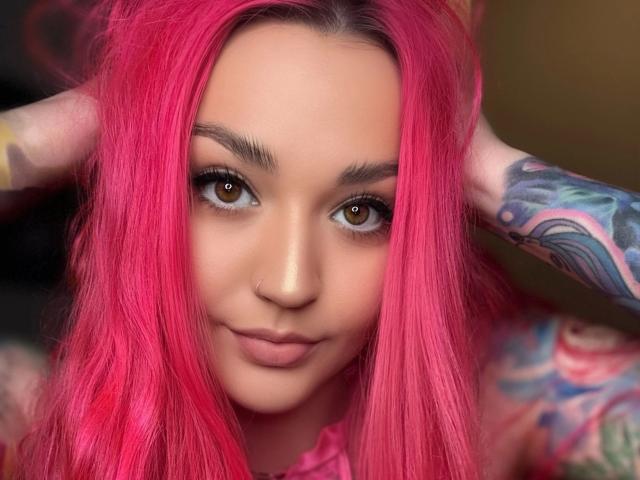PinkHurricane-hot with bright pink hair, tattooed arms, brown eyes, and a subtle smile, touching her head with hands. Her nose is pierced. She's looking directly at the camera.
