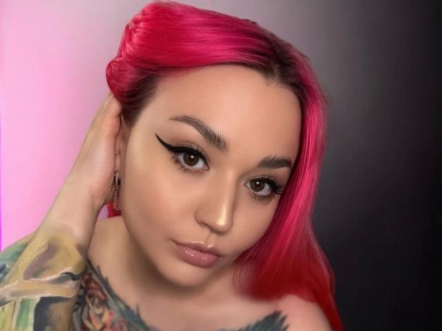 PinkHurricane-hot with bright pink hair, sharp black eyeliner, tattoos on her shoulder, and a subtle smile, touching her head. Her brown eyes and glossy lips are prominent.