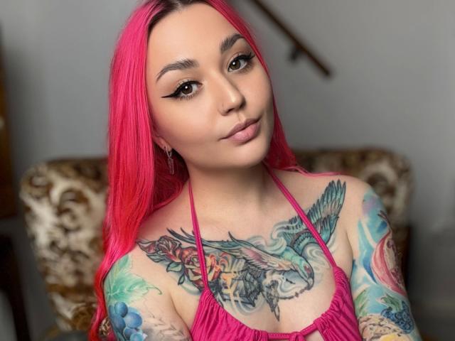 PinkHurricane-hot with bright pink hair, tattoos on chest and arms, wearing a pink halter top, looking at camera with winged eyeliner and pink lips.