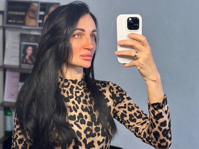TinaMart with long black hair, wearing a leopard print top, taking a selfie with a white phone, pink lips, ring on her left hand, and blue eyes.