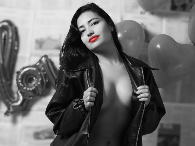 SilvannaReyes with dark hair, red lipstick, and black leather jacket, open to reveal her tits, holding balloons, in a black-and-white photo with one color, her lips.